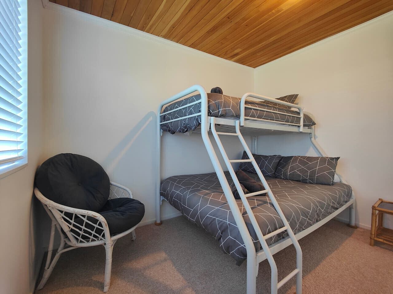 Un lit superposé avec une échelle, une chaise en osier blanche et une petite table en bois dans une pièce aux murs blancs et au plafond en bois.