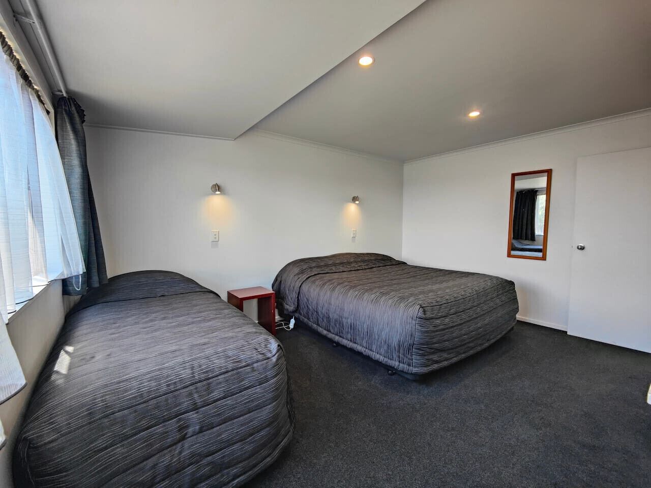 A small, single-room hotel room with two beds, a small wooden nightstand, a mirror, and a window with curtains.