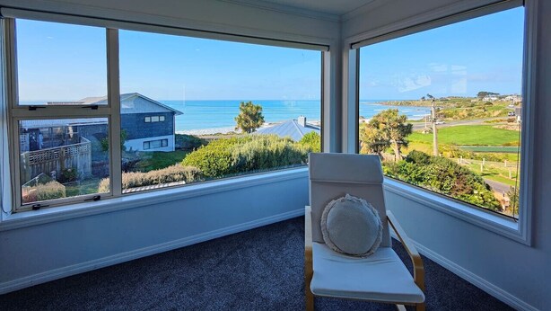 A room with large windows offering a view of the sea and a house across the water. - Riverton Helm`s Deep (Riverton / Aparima)