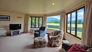 A spacious living room with a flat-screen TV, a comfortable sofa, a chair, and a large window offering a view of a green landscape. - Te Anau Dusky Delight (Te Anau)