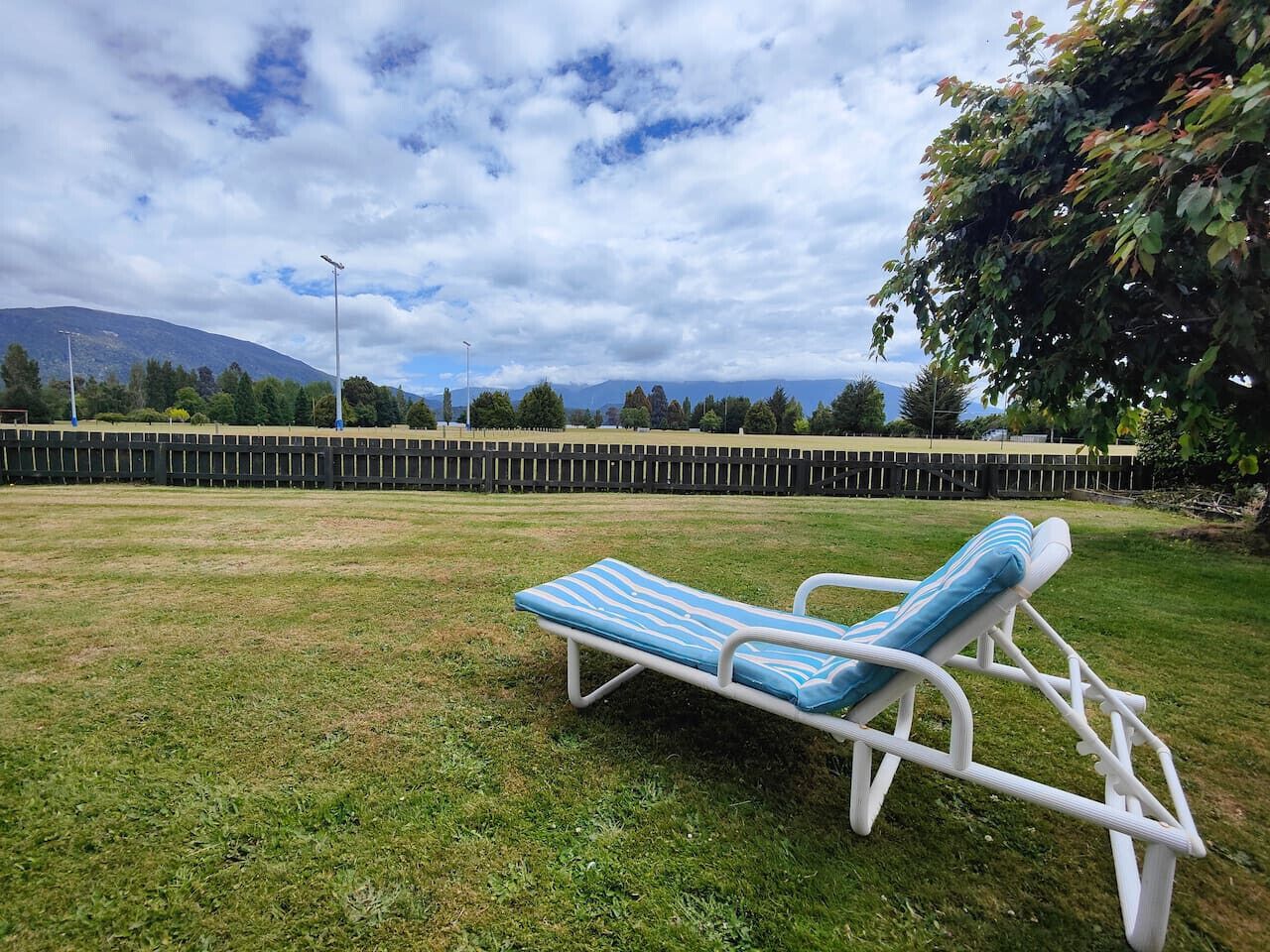 A single lounge chair with a striped cushion in a grassy area with a view of a mountain range and a cloudy sky.
