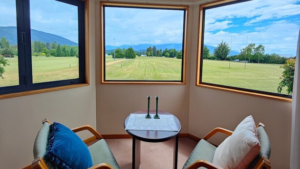 A corner room with three large windows offering a view of a grassy field and distant hills. There is a small table with two candles in the center. - Te Anau Dusky Delight (Te Anau)