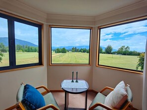 Habitación de esquina con tres ventanas amplias que dan a un campo verde y colinas lejanas. En el centro, hay una pequeña mesa con dos velas.