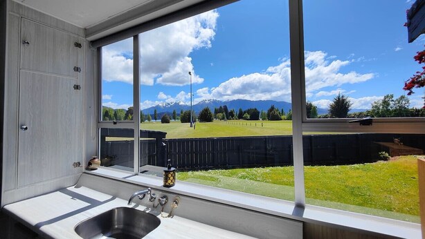 A kitchen with a large window, a sink, and a view of a grassy yard and mountains. - Te Anau Retro Retreat (Te Anau)