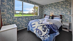 A bedroom with a large bed, a nightstand, a bedside table, and a window with a view of a green landscape. - Te Anau Retro Retreat (Te Anau)