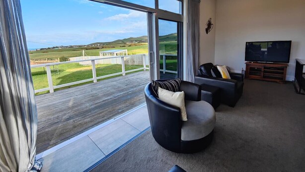 Een ruime woonkamer met een groot raam dat uitzicht geeft op een groen landschap. Er staat een zwarte leren bank met kussens en een flatscreen televisie op een houten dressoir.