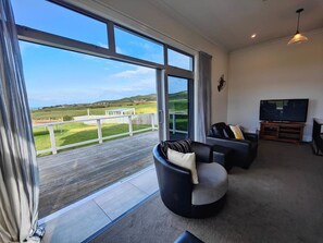 A spacious living room with a large window offering a view of a green landscape. There is a black leather sofa with cushions and a flat-screen TV on a wooden stand. - Church Street Charmer (Riverton / Aparima)