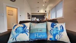 Een woonkamer met een zwarte leren bank, drie decoratieve kussens met blauwe krabmotieven, een wandklok en een keuken op de achtergrond.
