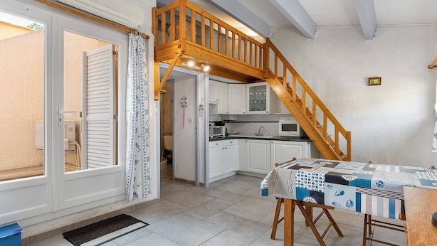 A compact kitchen with white cabinets, a wooden staircase, and a dining area with a table set for two. - STUDIO MEZZANINE CLIMATISE POUR 4 PERSONNES (LE CAP D AGDE)