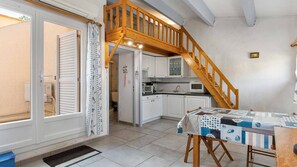 A compact kitchen with white cabinets, a wooden staircase, and a dining area with a table set for two. - STUDIO MEZZANINE CLIMATISE POUR 4 PERSONNES (LE CAP D AGDE)
