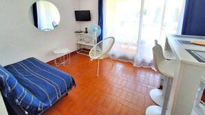 A room with a bed, a desk, a chair, a TV, and a window with curtains. - STUDIO CABINE CLIMATISE AVEC CHAMBRE ALCÔVE (LE CAP D AGDE)