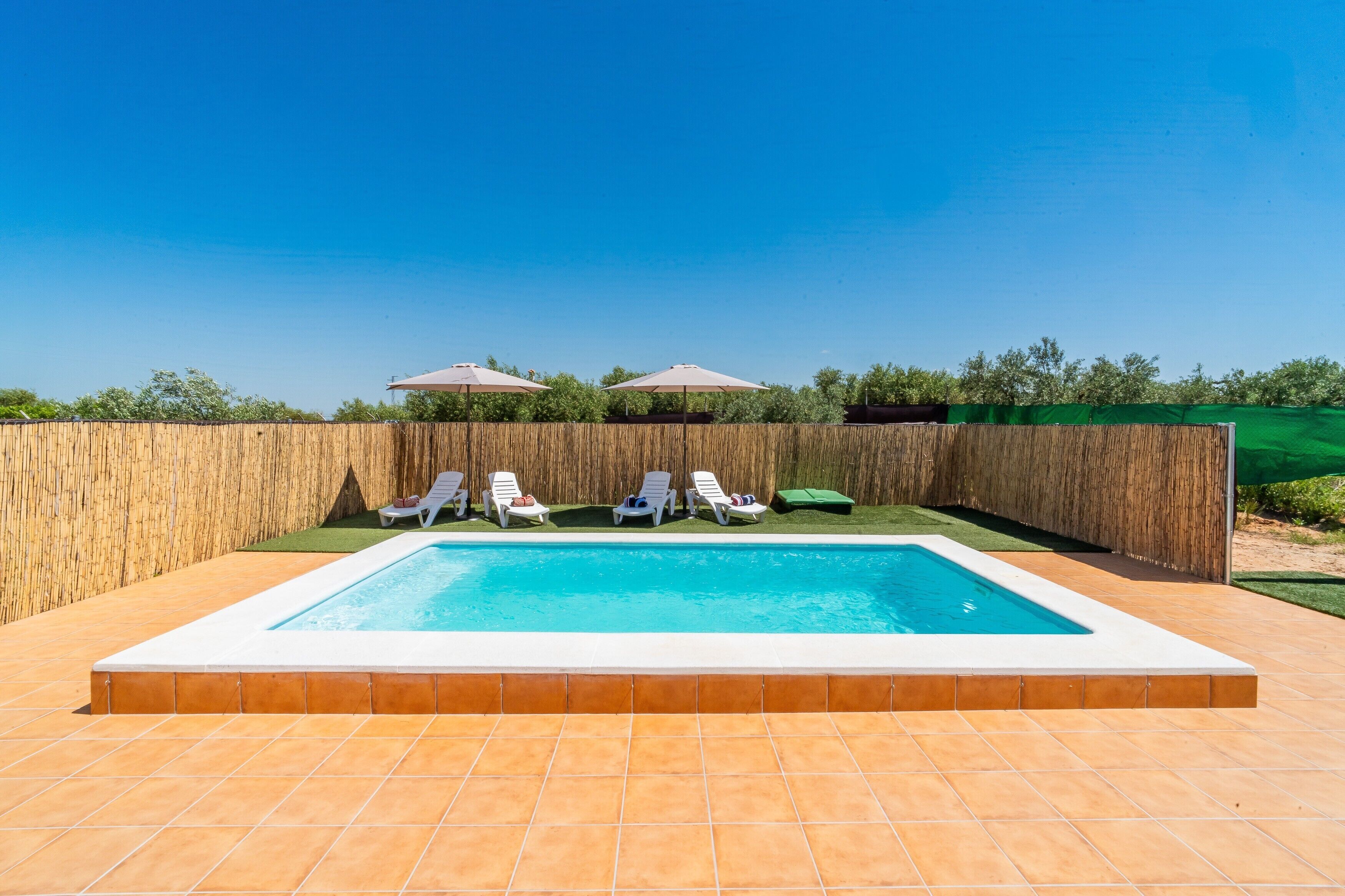 Una piscina rectangular con borde de azulejos, rodeada por una valla de madera y equipada con varias sillas de descanso y sombrillas.