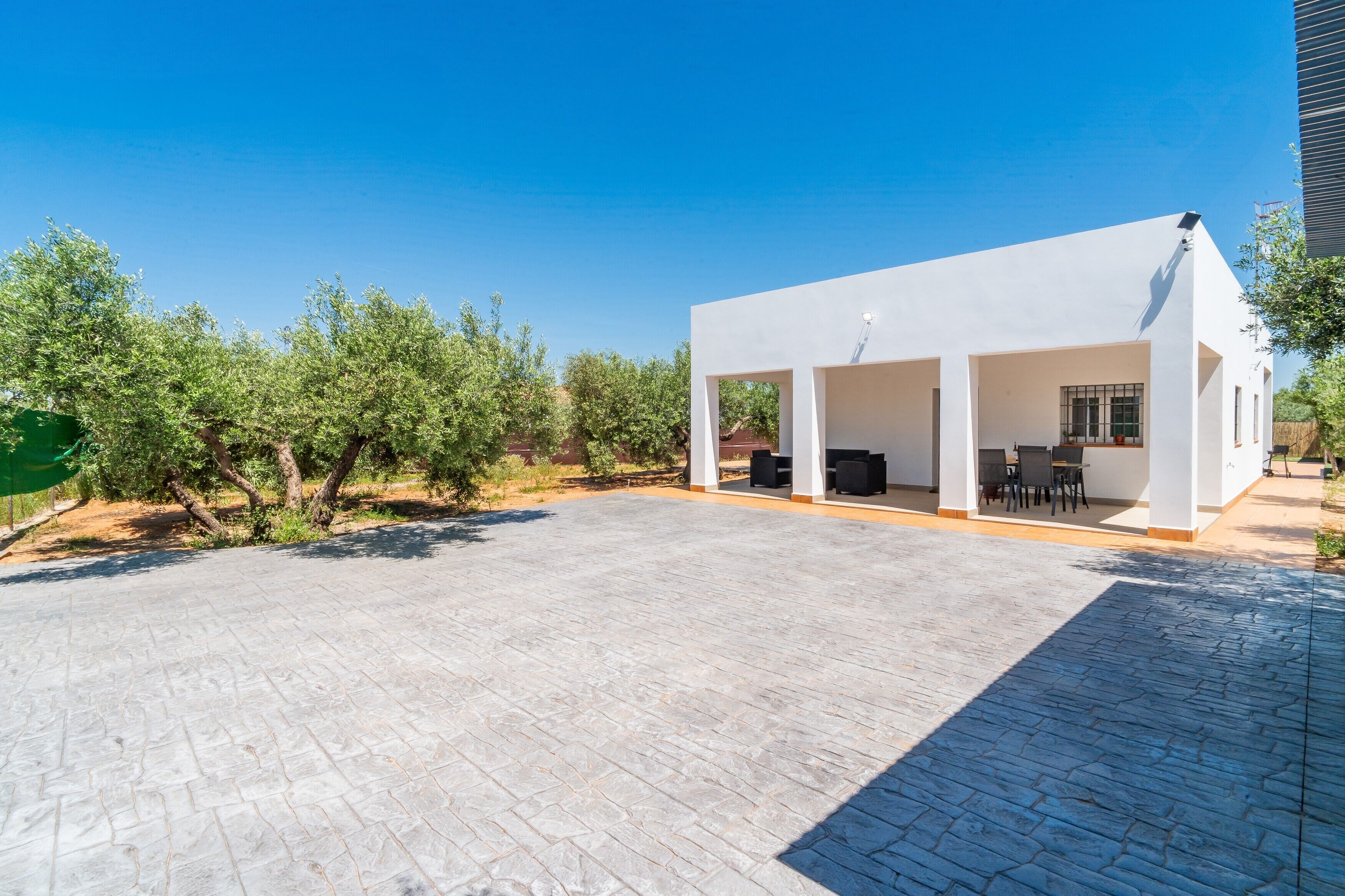 Un patio al aire libre moderno con una estructura blanca, piso empedrado y abundante vegetación.