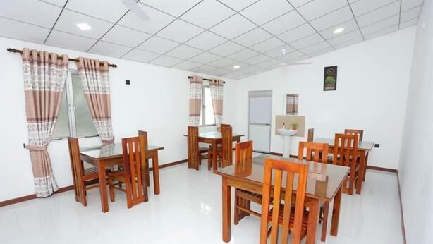 A dining area with wooden tables and chairs, a white tiled floor, and curtains on the windows. - Kumbuk Sewana Villa (Anuradapura)