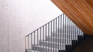 Una escalera con barandilla de vidrio, paredes de concreto y techo inclinado de madera.