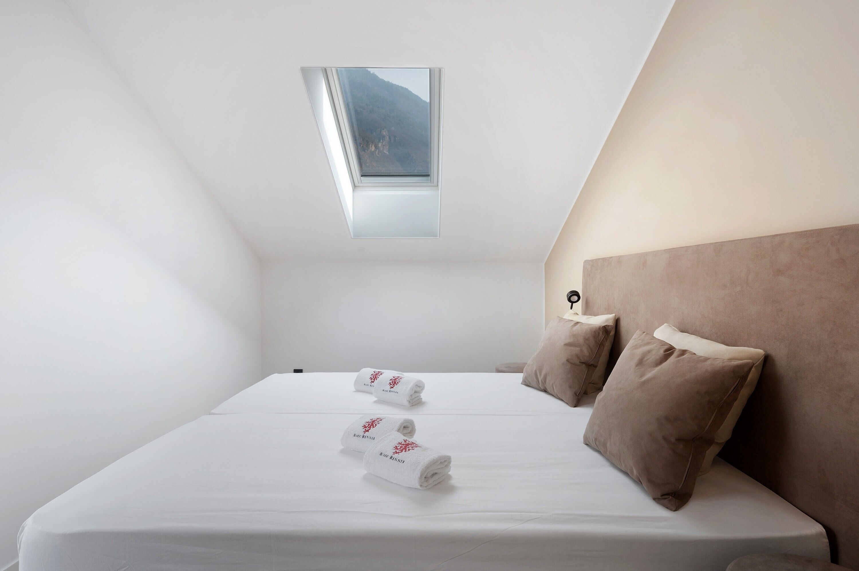 Une chambre avec un puits de lumière, un lit avec du linge de lit blanc, deux oreillers et une table de chevet.