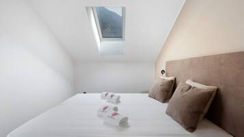 Une chambre avec un puits de lumière, un lit avec du linge de lit blanc, deux oreillers et une table de chevet.