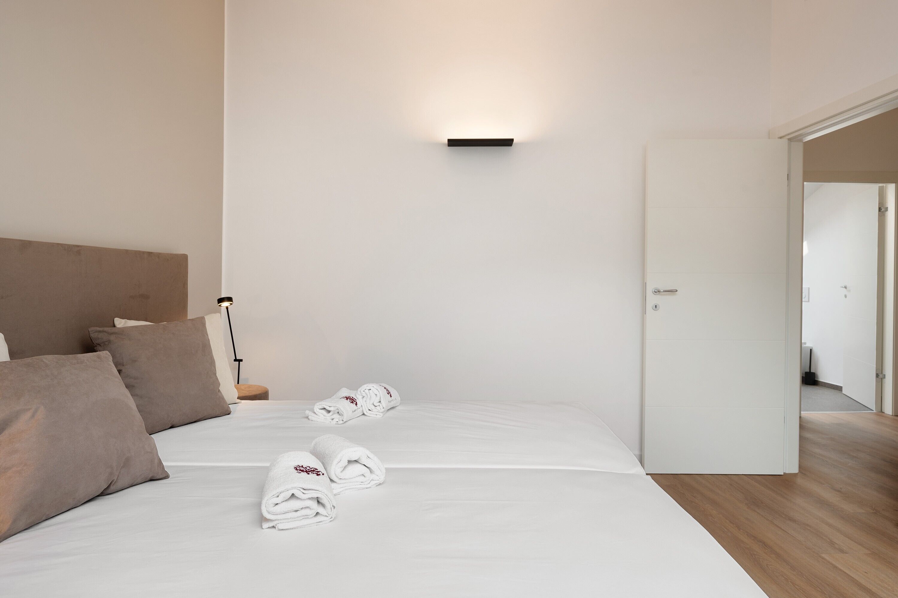 A hotel room with a bed, pillows, a nightstand, a wall-mounted light, and a door.