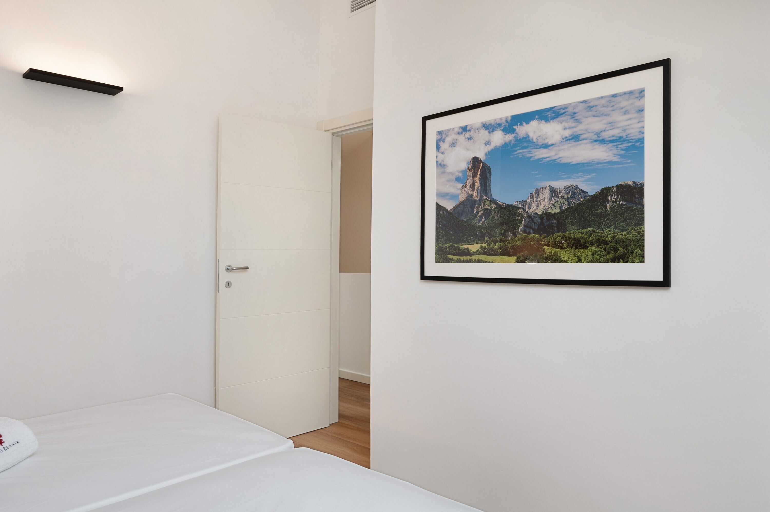 Hotelkamer met een bed, een lijst met een landschapsfoto, een deur naar een andere kamer en een klein plankje met een lampje.