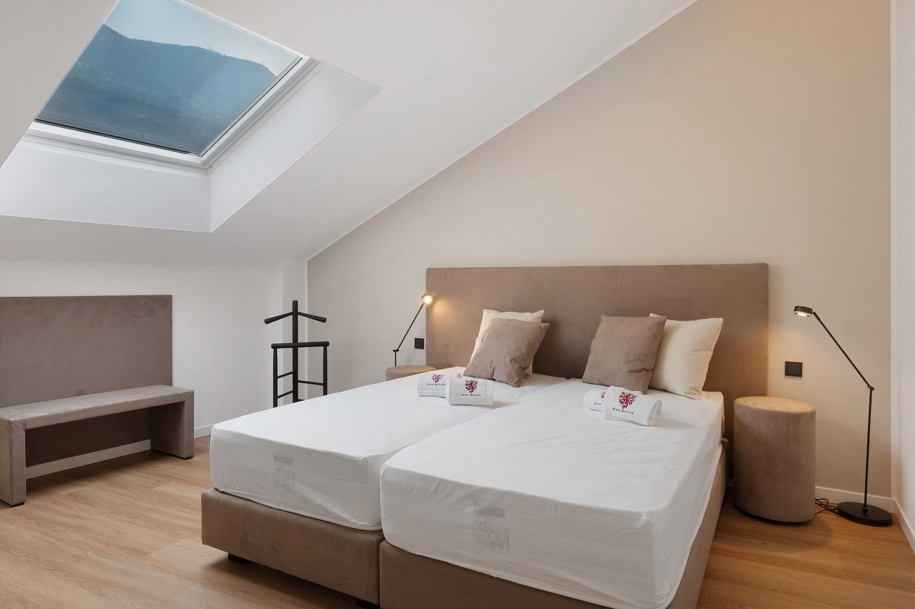 A bedroom with a skylight, a bed with two pillows, a bedside lamp, and a bench.