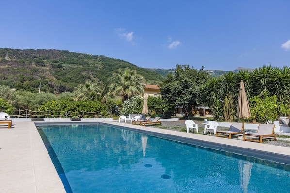 A swimming pool with lounge chairs and umbrellas, surrounded by greenery and hills.
