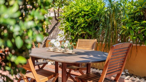 Une table en bois placée à l’extérieur, avec des chaises, entourée de verdure et posée sur un sol carrelé.