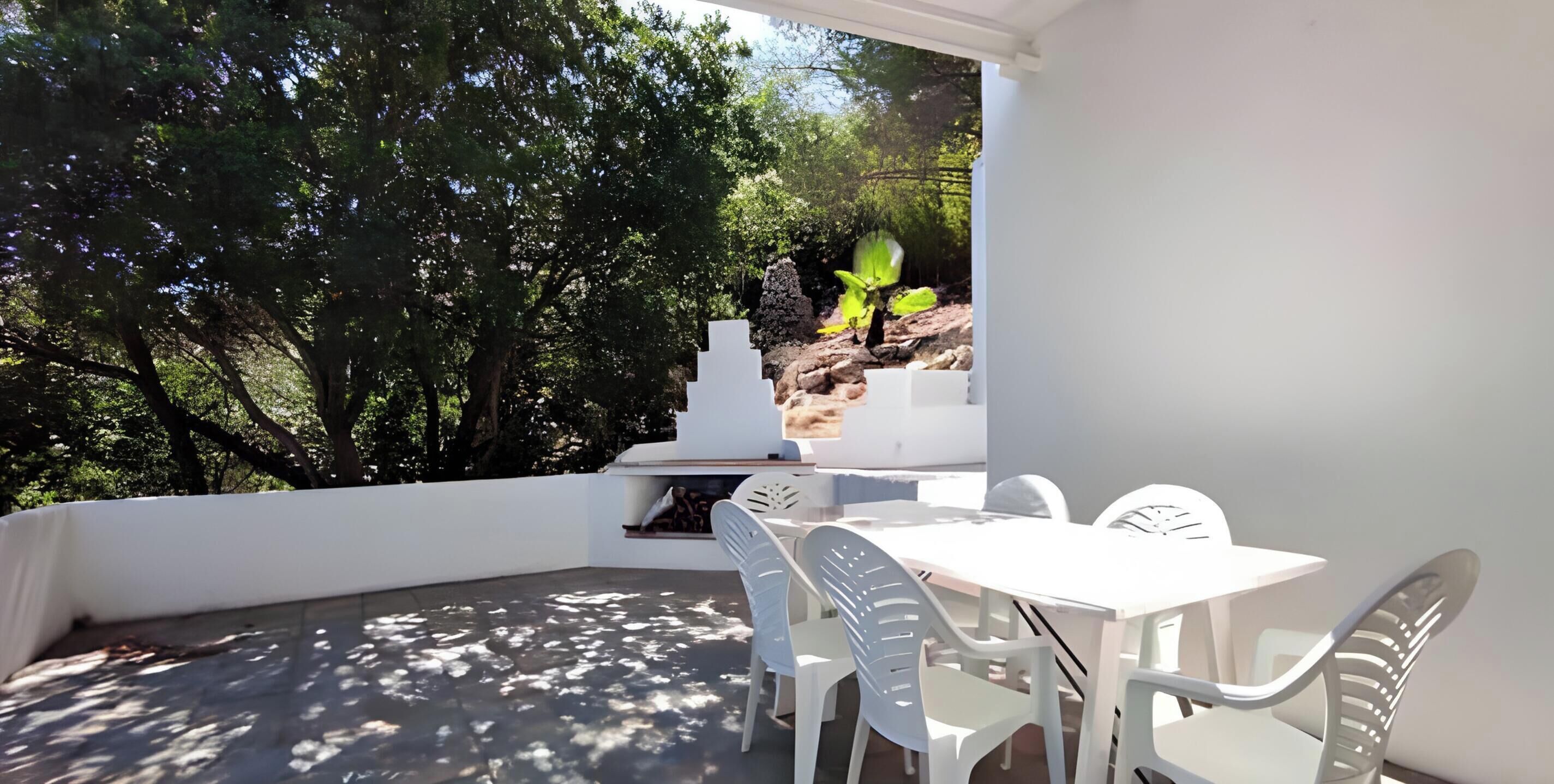 Um pátio com mesa e cadeiras, uma parede branca e vista para árvores e uma formação rochosa.