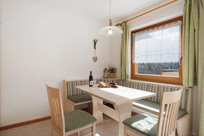 Un espace repas comprenant une table et des bancs, un luminaire suspendu, une fenêtre avec des rideaux et une vue sur l’extérieur.