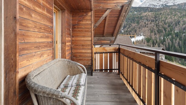 Ein Holzbalkon mit einem Korbstuhl, Holzwerteln und Blick auf die Berge.