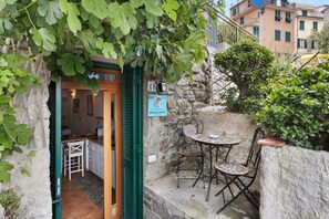 Un mur de pierre avec une porte, un petit espace extérieur aménagé avec des chaises en métal, et une enseigne indiquant le numéro de l’immeuble.