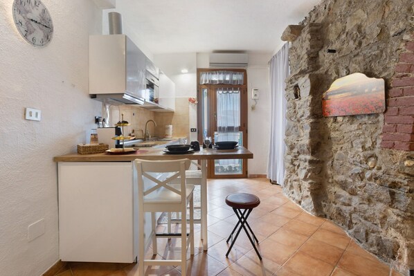 Une cuisine avec un réfrigérateur blanc, un îlot central en bois, une chaise blanche, un petit tabouret et un mur en pierre.