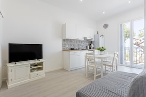 Ein modernes, helles Zimmer mit einem Flachbildfernseher, einer Essecke mit Tisch und Stühlen, einer Küche mit weißen Schränken und einem Balkon mit Aussicht.