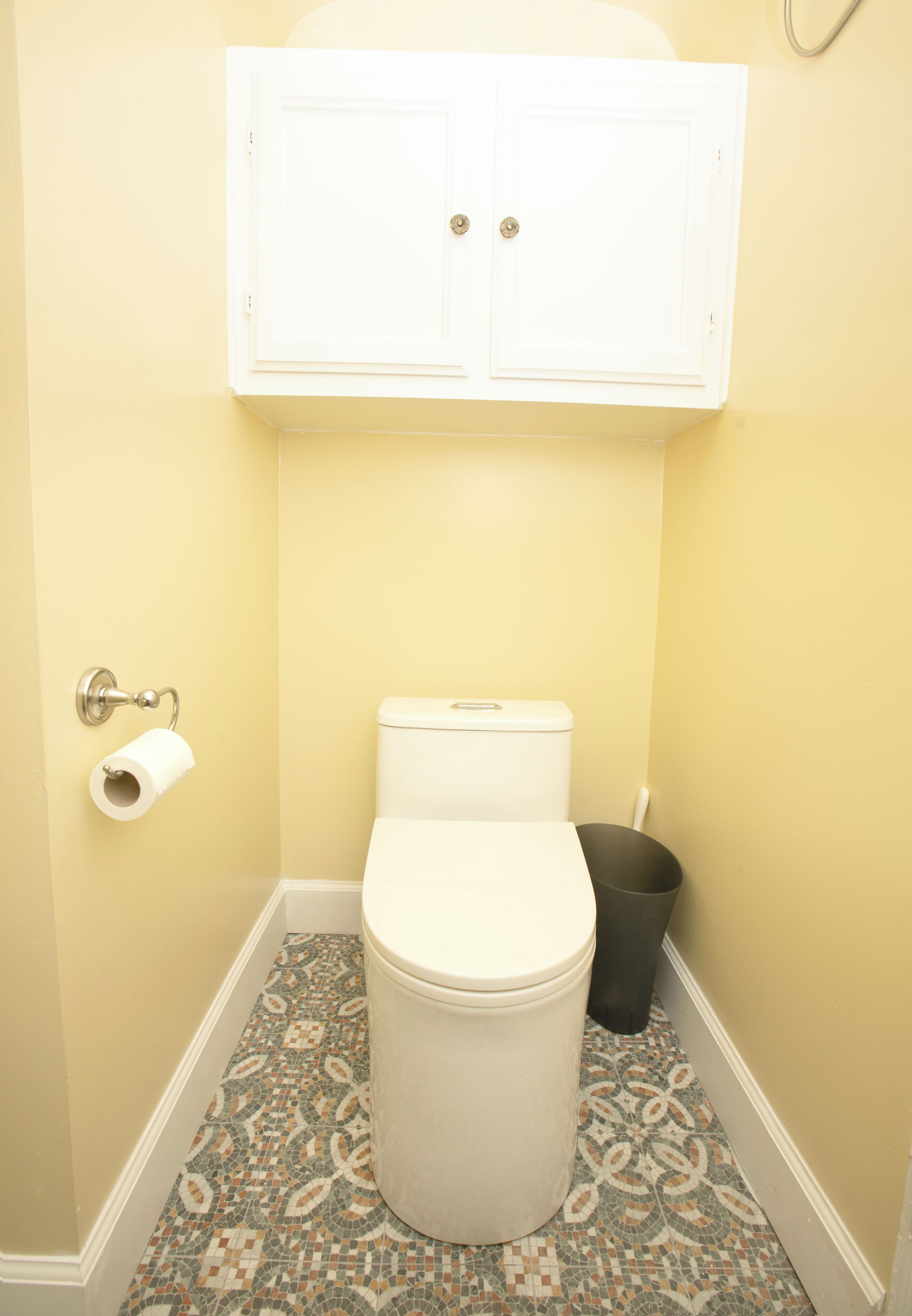 Un baño pequeño con inodoro, un dispensador de papel higiénico, un cubo de basura y un armario sobre el inodoro.