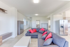 A modern living room with a grey sectional sofa, a white coffee table, and a kitchen area with a stainless steel refrigerator.