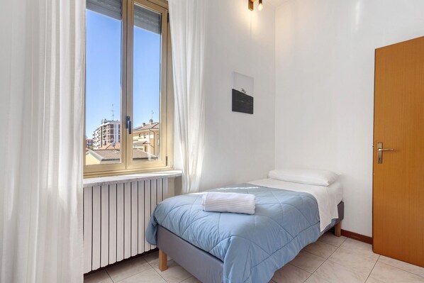 A bedroom with a bed, a window with curtains, a radiator, a door, and a wall-mounted picture.