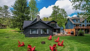 Une cabane en rondins avec une cheminée en pierre, une terrasse dotée de meubles d’extérieur et un foyer entouré de fauteuils Adirondack rouges.