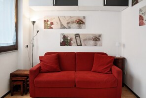 A red sofa in a room with a white wall, a small wooden table, and two wall-mounted artworks depicting Paris and London.