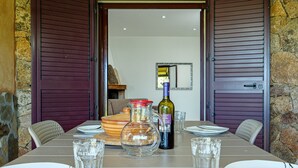 Une table à manger dressée avec des assiettes, des verres, une bouteille de vin et un pichet, avec un mur en pierre et des volets en bois en arrière-plan.