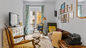 Una sala de estar con televisión, máquina de escribir, sofá, sillón, mesa y ventana con cortinas.