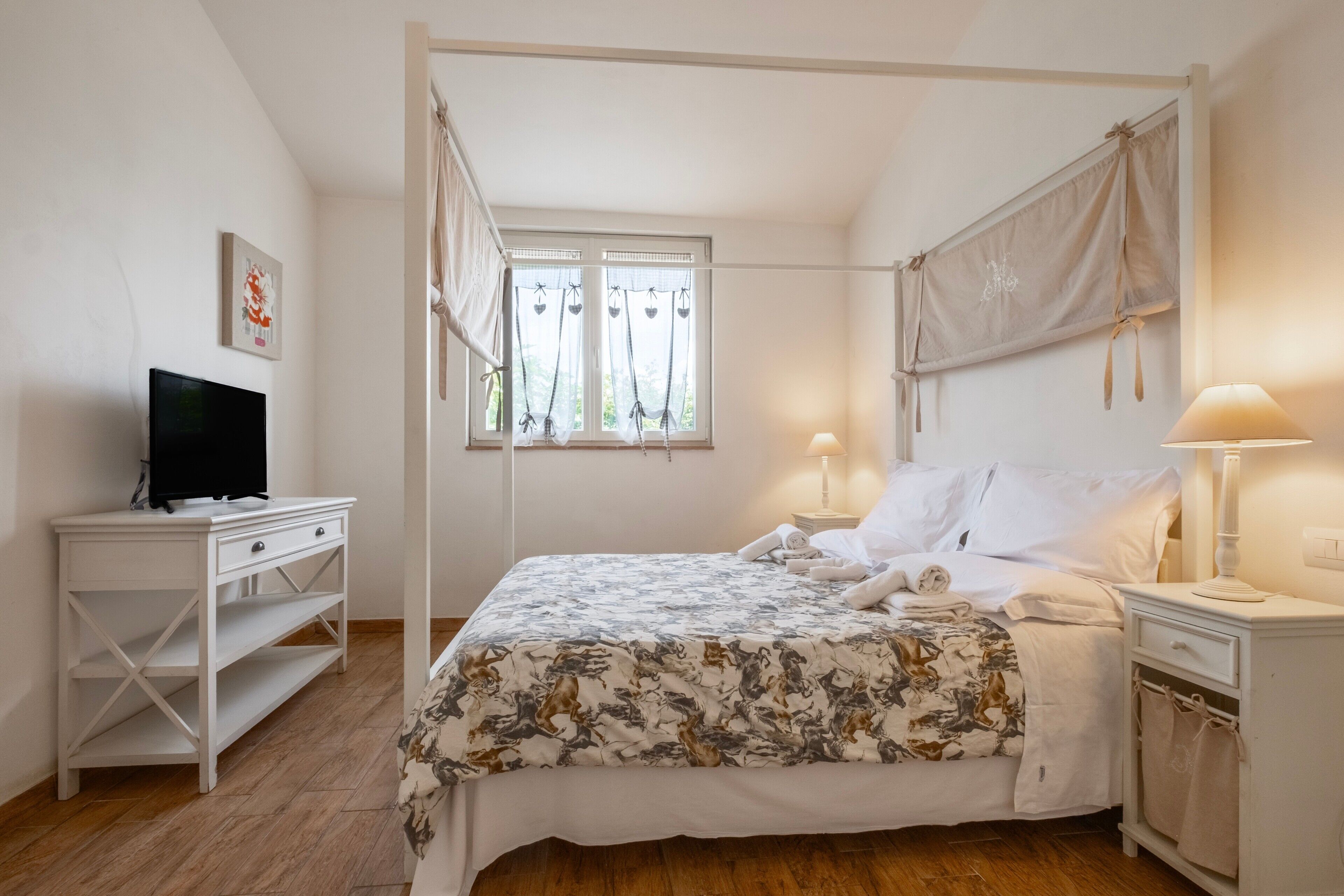 Ein Schlafzimmer mit Himmelbett, einem Fernseher auf einer weißen Kommode und einem Fenster mit Vorhängen.