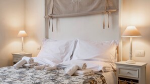 Un lit bien fait, avec du linge de lit blanc et des serviettes pliées, deux lampes de chevet et un simple luminaire fixé au mur.
