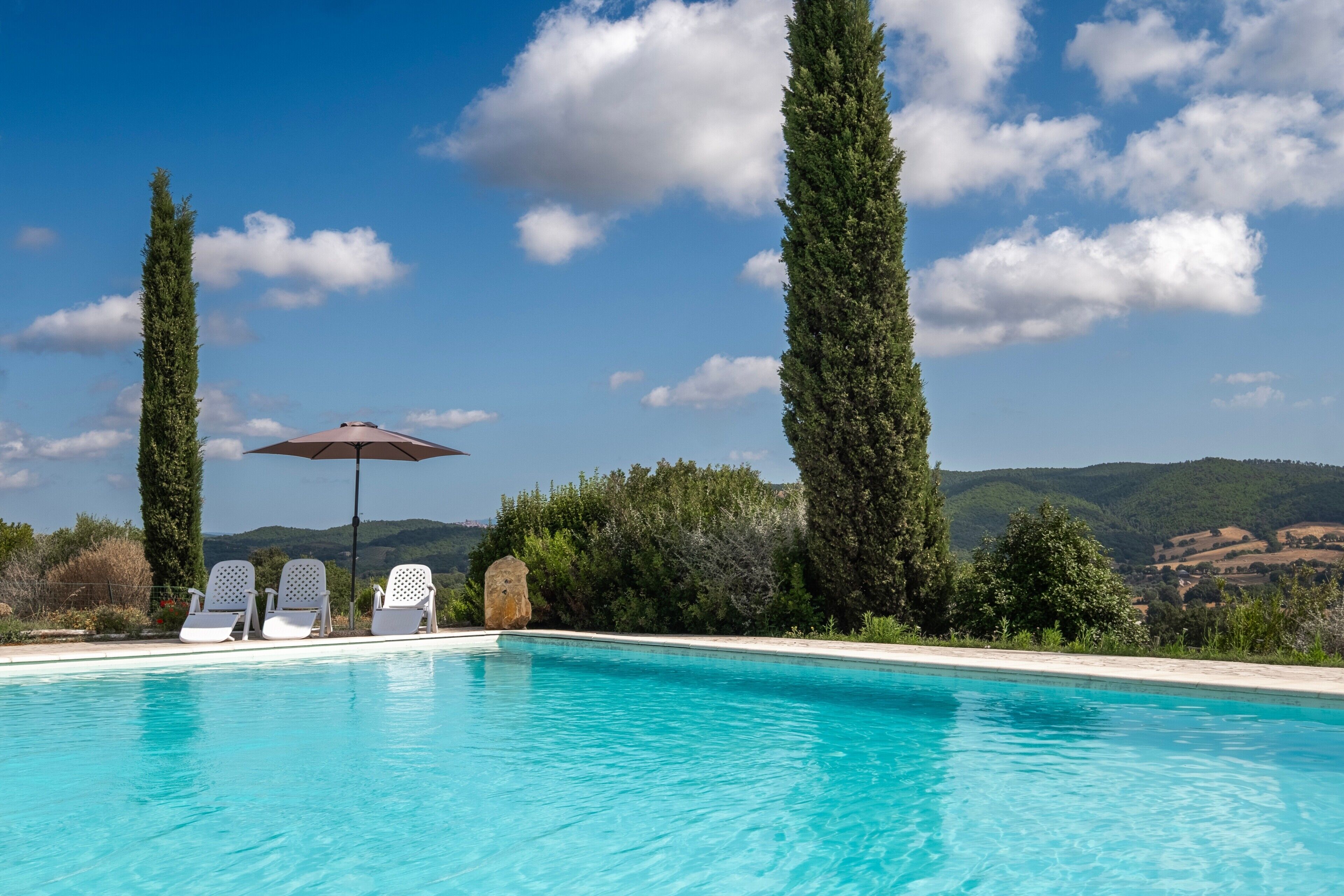Una piscina con sillones de descanso y una sombrilla, rodeada de altos árboles y un paisaje montañoso.