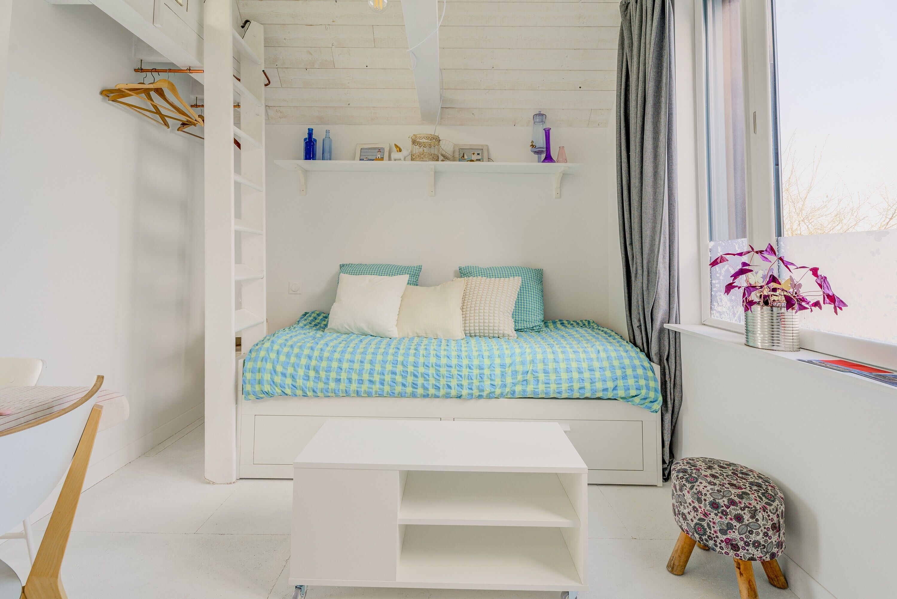 Une chambre à coucher comprenant un lit, une petite table, une chaise, une étagère avec quelques objets et une fenêtre avec une plante.