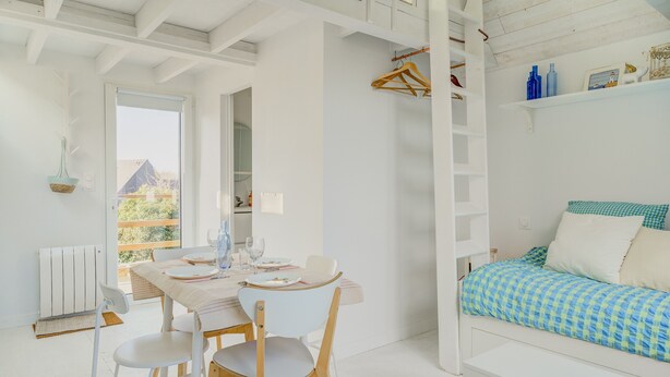 Una habitación luminosa con una cama, una mesa con sillas, una percha y un radiador.