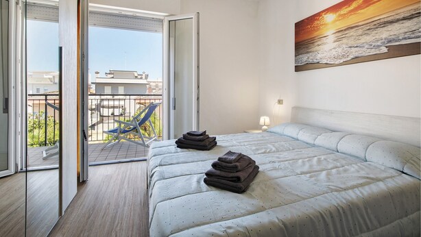A hotel room with a bed, a painting of a sunset, and a balcony with a chair.