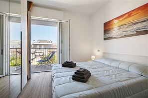 A hotel room with a bed, a painting of a sunset, and a balcony with a chair.
