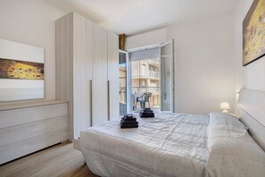 Une chambre à coucher avec un lit, une armoire, un tableau et un balcon donnant sur des immeubles.