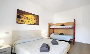 A hotel room with a single bed, a bunk bed, a painting on the wall, and a bedside table with a lamp.
