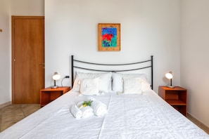Een keurig opgemaakt bed met witte lakens en opgerolde handdoeken, een houten hoofdbord, een schilderij boven het bed, en twee nachtkastjes met lampen.