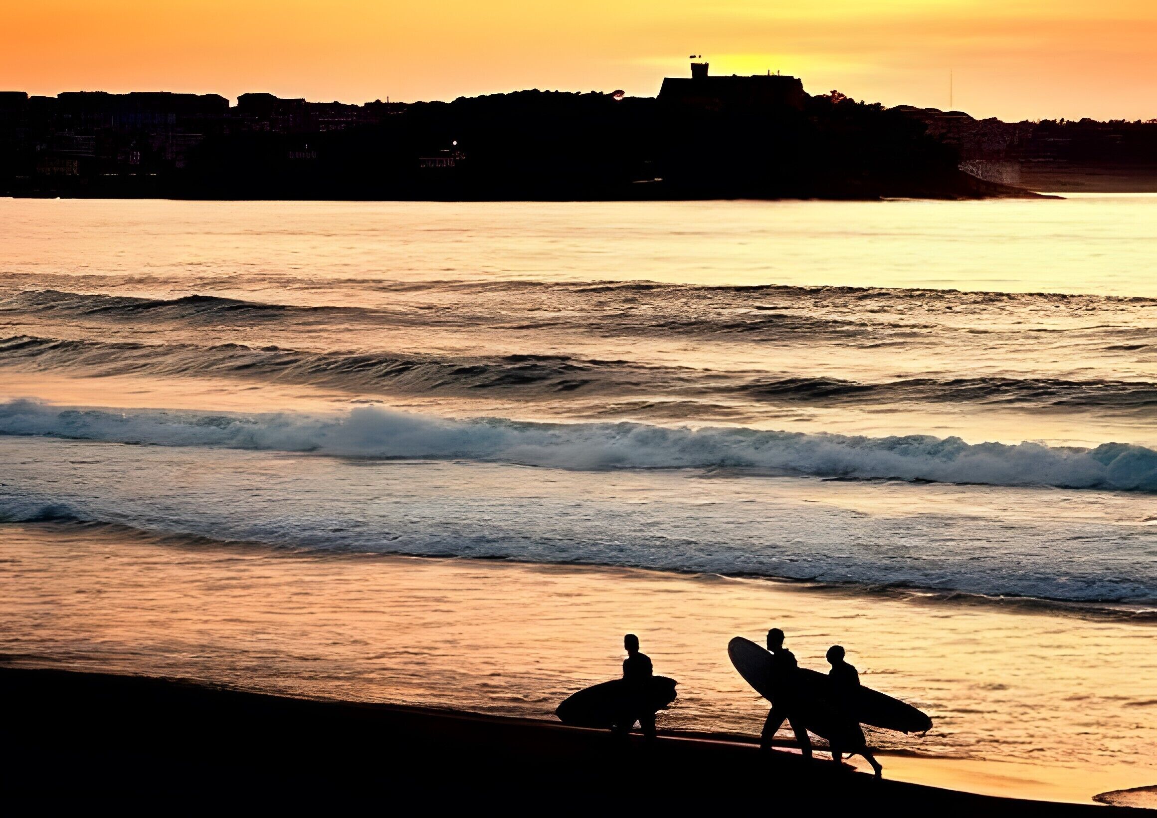 Escena de playa con olas y surfistas silueteados contra el atardecer.